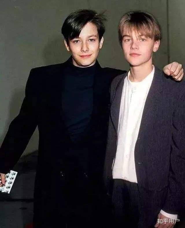 two young men standing next to each other in front of a white wall and one is wearing a black suit