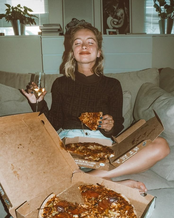 a woman sitting on a couch with two pizzas in front of her and one holding a glass of wine