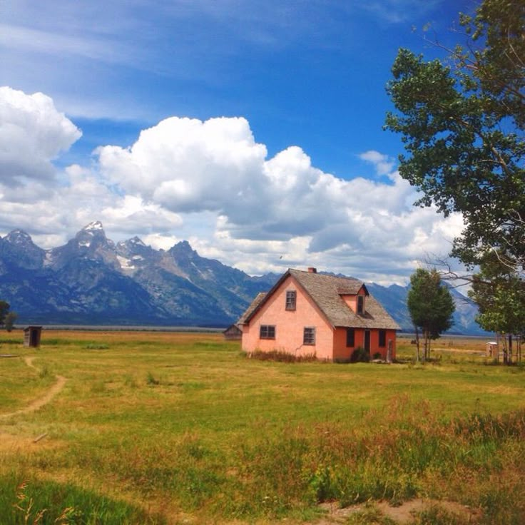 a small pink house sitting in the middle of a field with mountains in the background