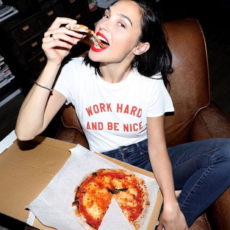 a woman sitting in a chair eating pizza from a box on her lap and holding a piece of pizza up to her mouth