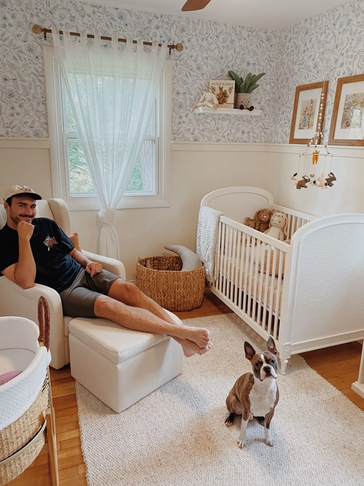 a man sitting in a white chair next to a small dog on the floor near a crib