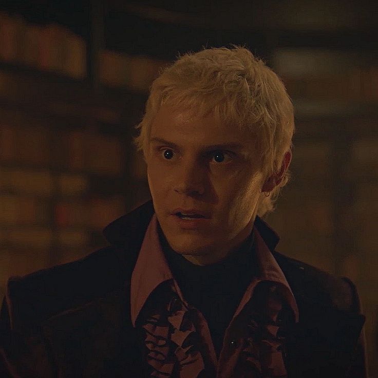 a man with blonde hair wearing a black jacket and pink shirt in a dark room