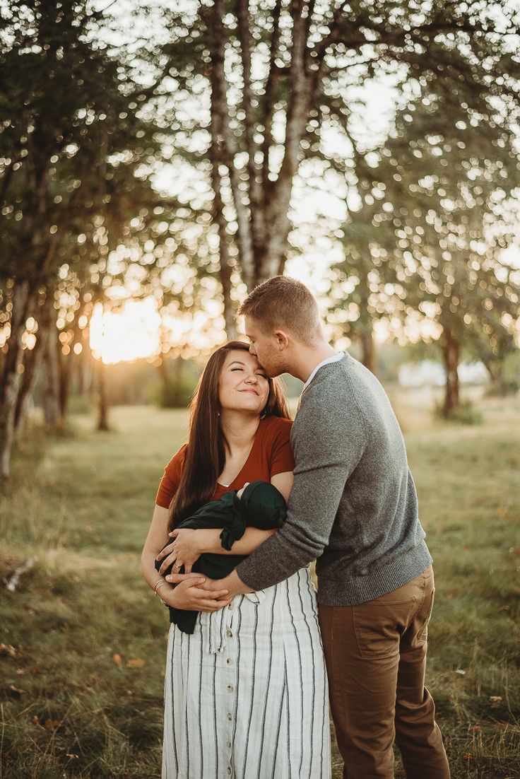 a man and woman embracing each other while standing in front of trees with the sun setting behind them