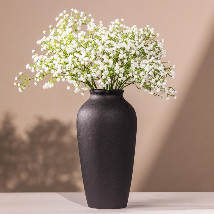 a black vase filled with white flowers sitting on top of a table next to a wall