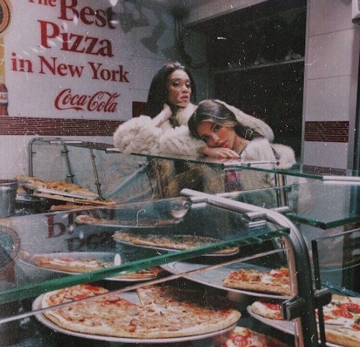 a man and woman standing in front of a pizza counter with many different kinds of pizza on it