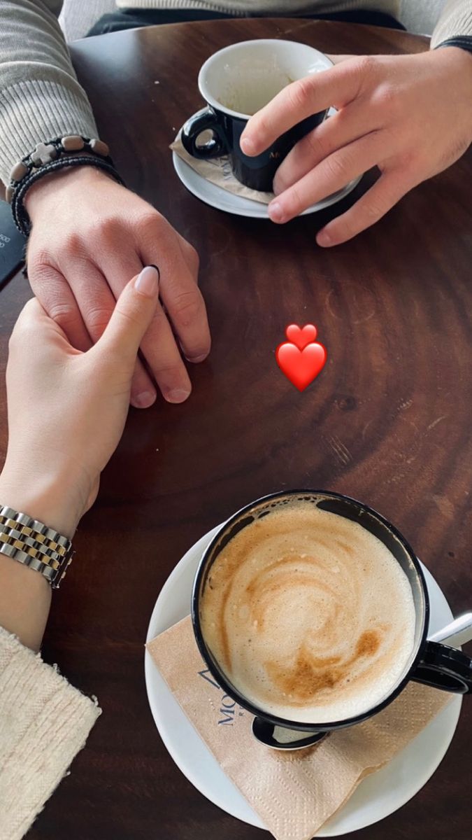 two people holding hands over a cup of coffee on a table with a red heart