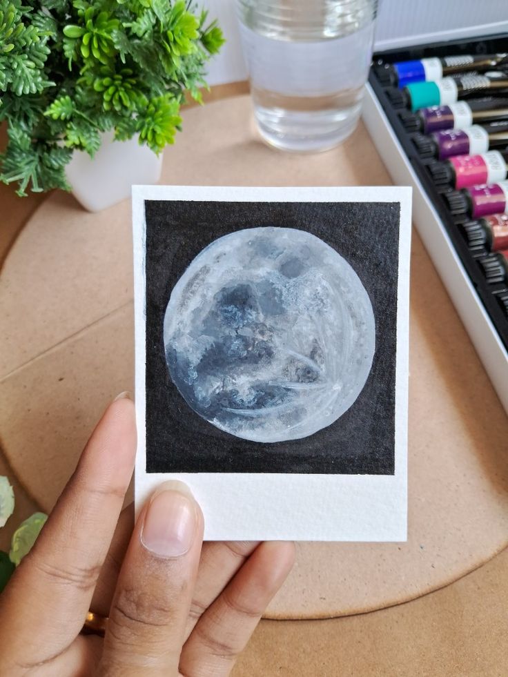 a person holding up a photo of the moon in their left hand, next to some watercolors