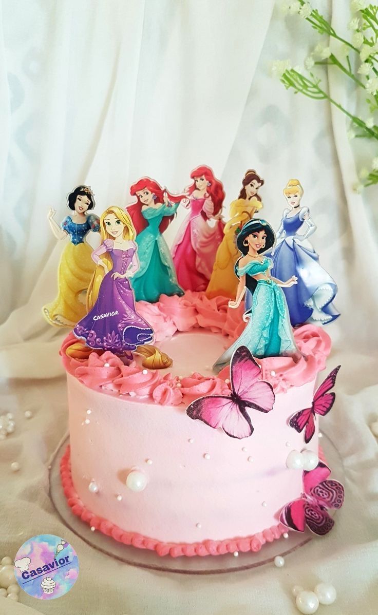 there is a pink cake with princess figures on it and flowers in the back ground