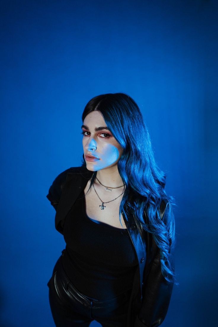 a woman with blue hair posing for a photo in front of a blue background and wearing black leather pants