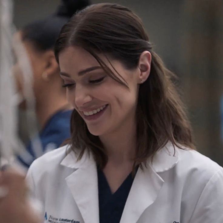 a woman in white lab coat smiling and looking at her cell phone while another person looks on