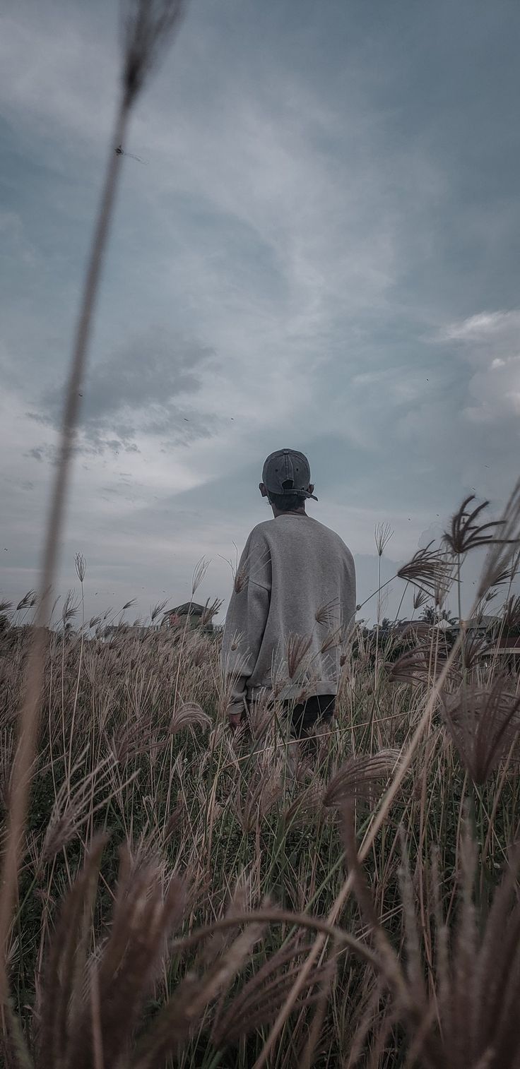 a man standing in the middle of a field with tall grass and cloudy sky behind him