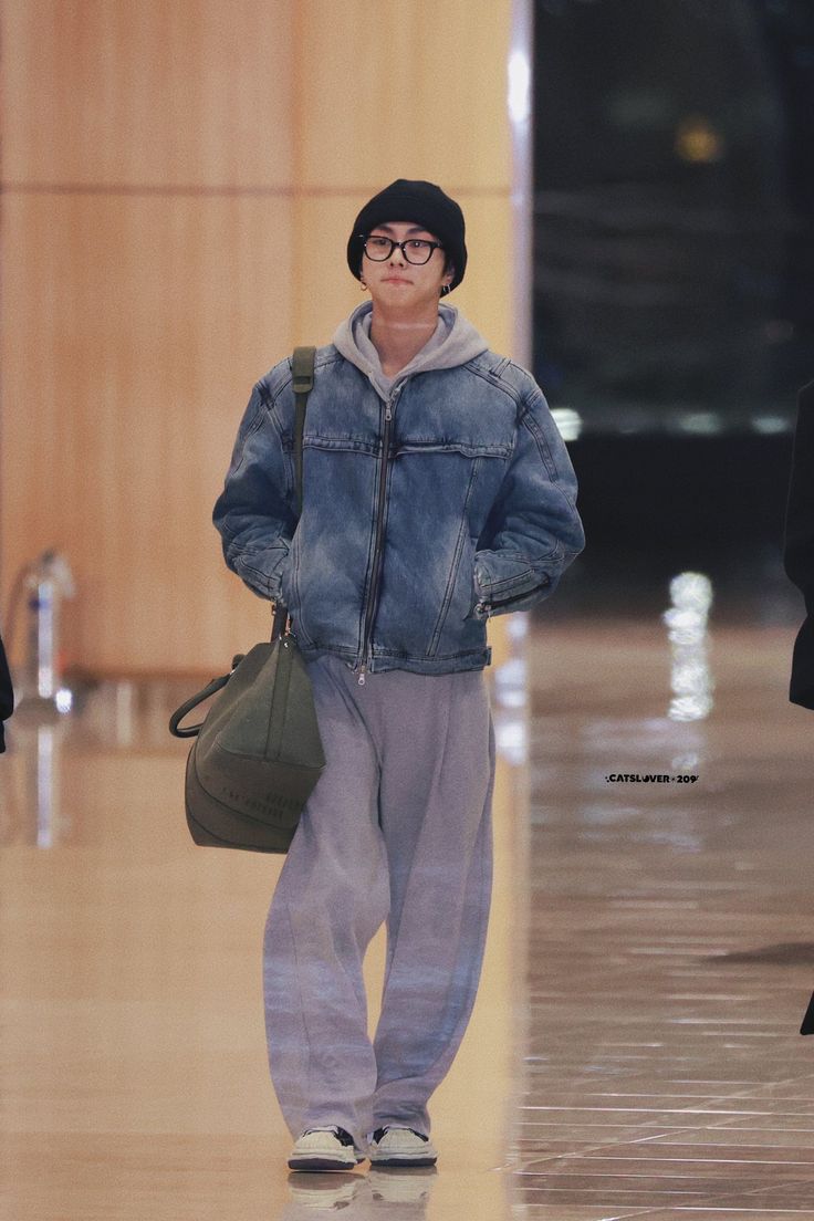 a man walking down a hall with a handbag on his shoulder and wearing a denim jacket