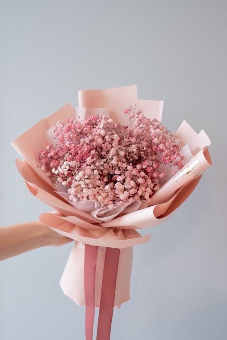 a person holding a bouquet of flowers with pink ribbons on the end and white wall behind it