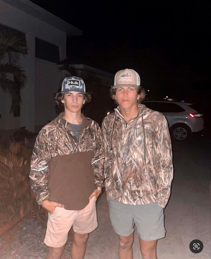 two young men standing next to each other in front of a house at night with the lights on