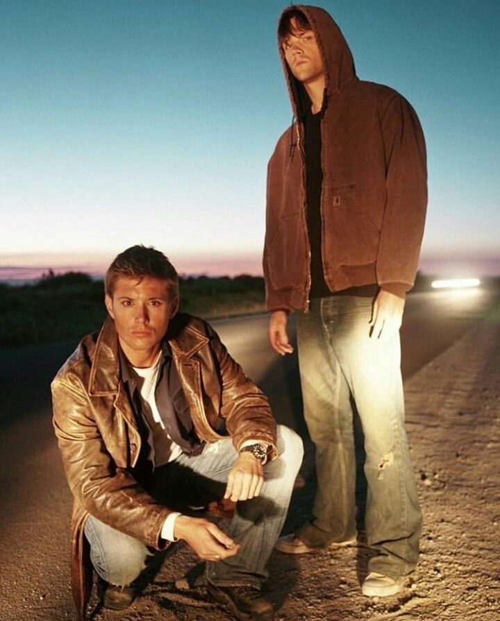 two men sitting on the side of a road next to each other, one wearing a brown jacket