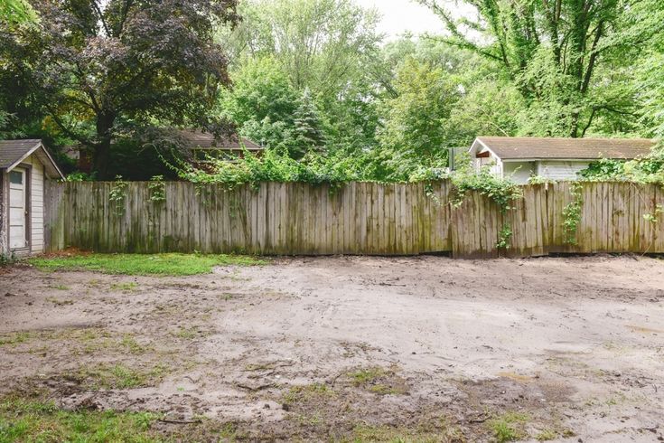 an empty yard next to a fence with trees in the back ground and bushes growing on it