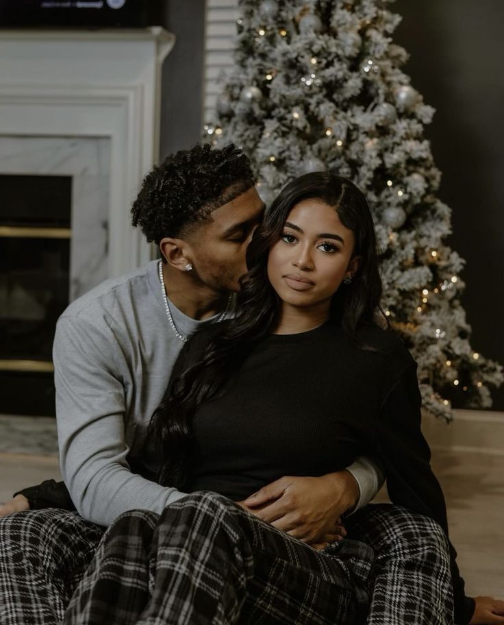 a man and woman sitting next to a christmas tree in front of a fire place