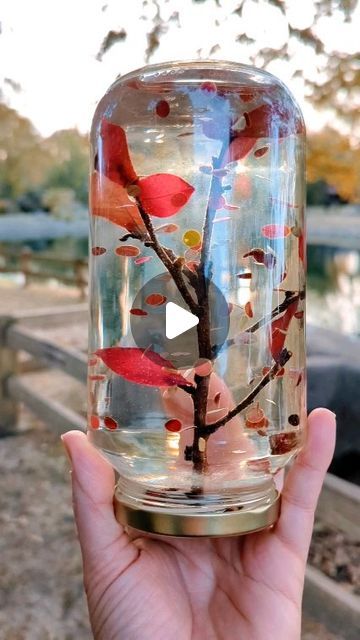 a person holding up a glass jar with a tree in it that has red leaves on it