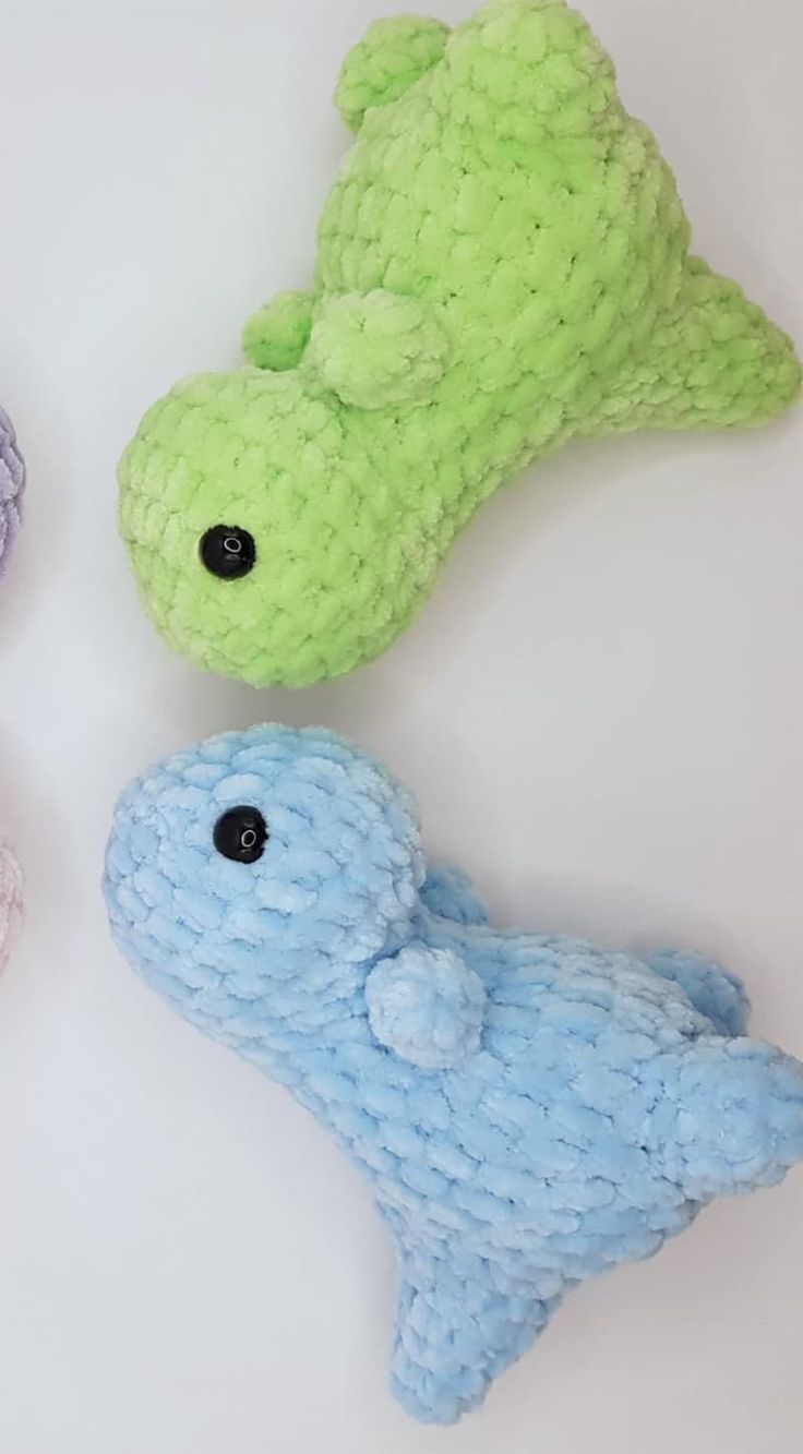 three stuffed animals sitting next to each other on a white surface, one is green and the other two are blue