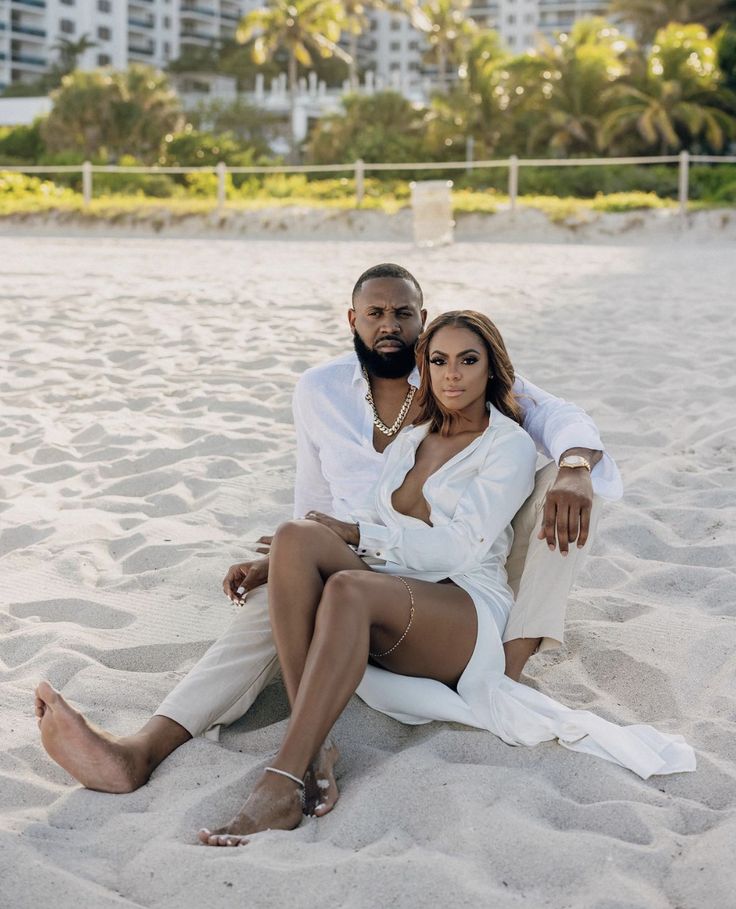 a man and woman sitting in the sand on a beach with palm trees behind them