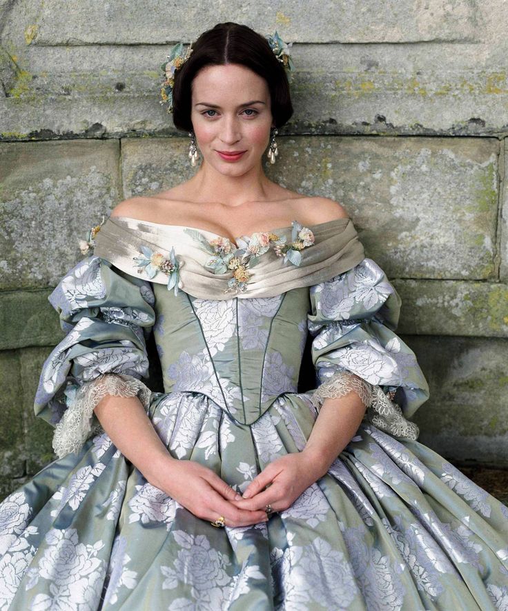 a woman in a green and white dress with flowers on her head, sitting against a stone wall