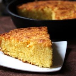 a piece of cornbread on a white plate next to a black skillet with another dish in the background