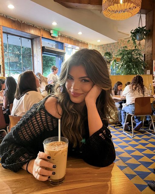 a woman sitting at a table with a drink in her hand and looking into the camera