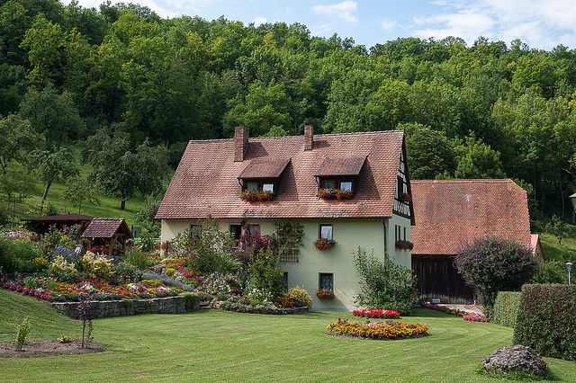 a house in the middle of a lush green field with lots of flowers around it