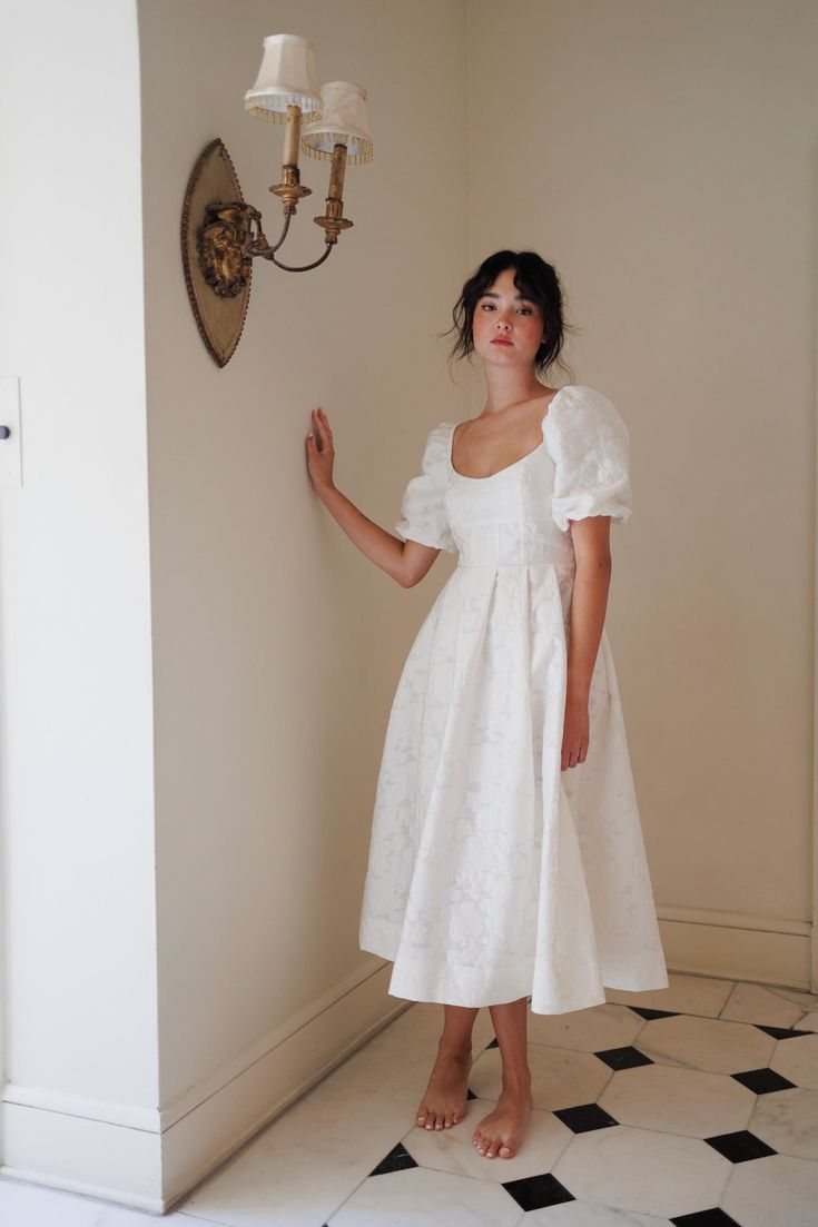 a woman in a white dress standing next to a wall with a clock on it