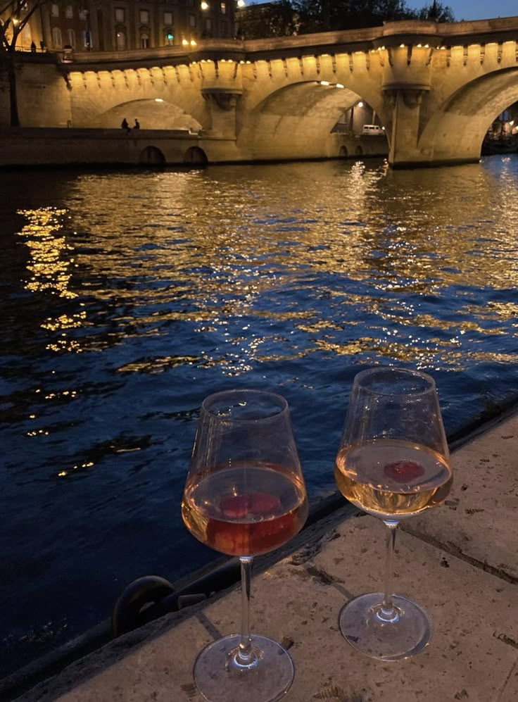 two glasses of wine are sitting on the edge of a ledge overlooking water and a bridge