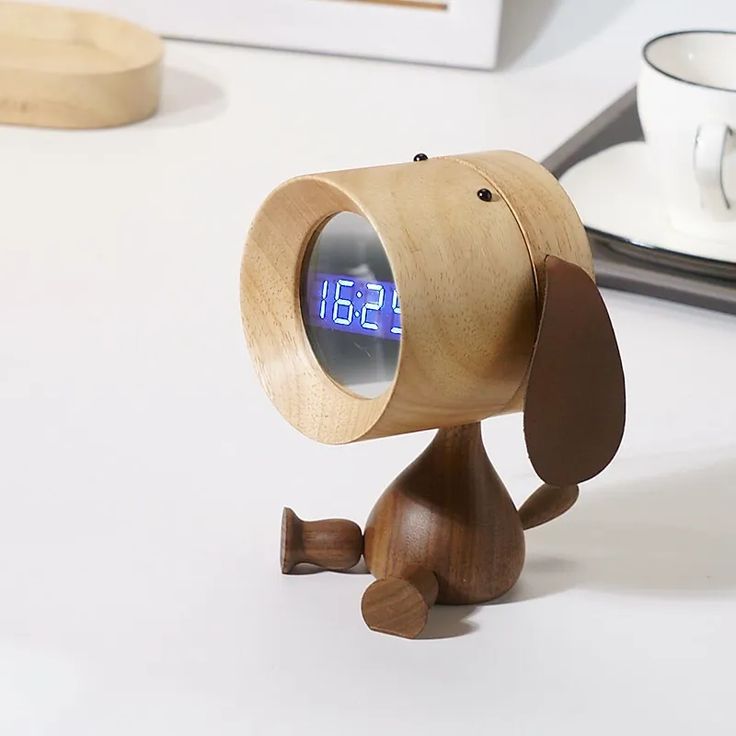 a wooden dog alarm clock sitting on top of a white table next to a cup