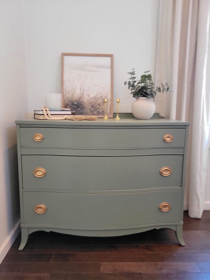 a green dresser with gold handles and knobs in a white walled room next to a window