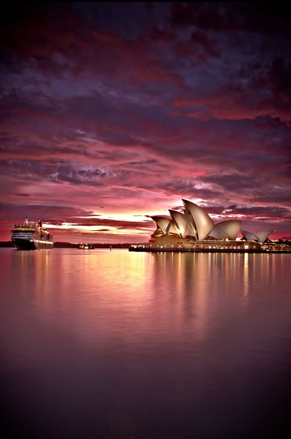 the sydney opera house is lit up at night with pink and purple clouds in the background