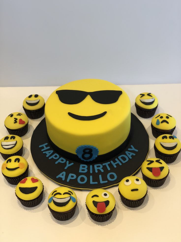 a birthday cake decorated with yellow and black smiley face cupcakes on a white table