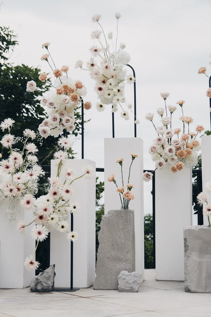 several white vases with flowers in them sitting on cement blocks near trees and bushes