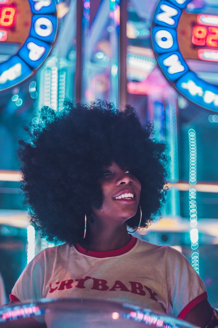 a woman with an afro smiles in front of neon signs and lights at the fairground