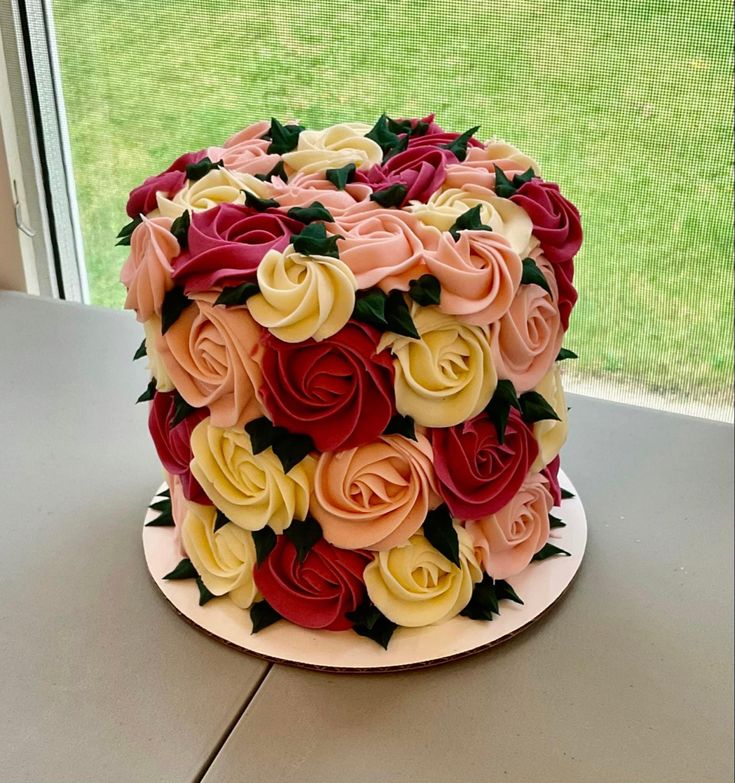 a multicolored cake with roses on it sitting on a table next to a window