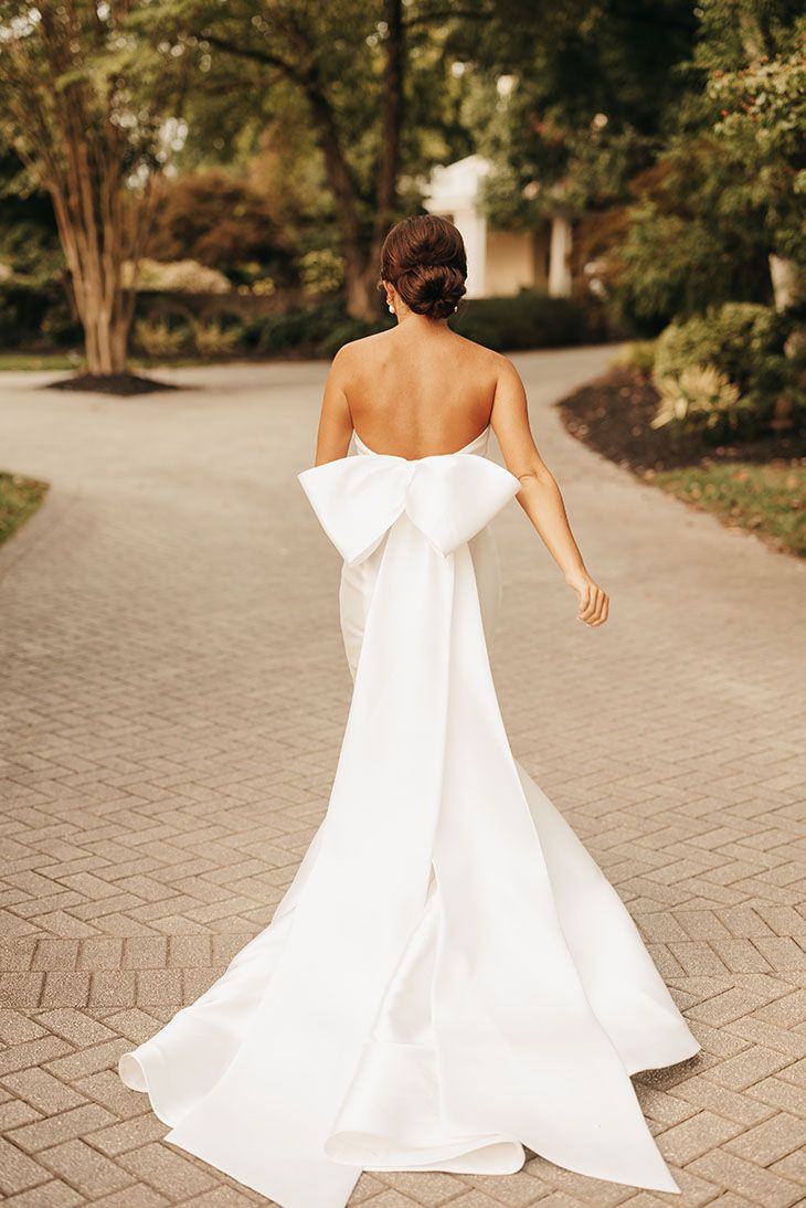 Classic and Elegant Summer Bride Style with a Full Neat Bridal Updo and Low Back Mermaid Fit Gown with Large Pressed Bow Wedding Dress With Back Out, White Wedding Dress With Bow, White Bow Wedding Dress, Ralph Lauren Bride, Strapless Timeless Wedding Dress, Wedding Gown Bow Back, Strapless Bow Wedding Dress, Mermaid Wedding Dress With Bow On Back, Spring Outfits 2024 Dress