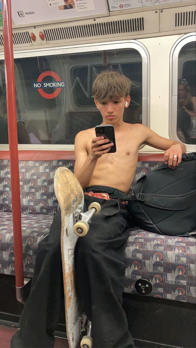 a shirtless man sitting on a train holding a skateboard and looking at his cell phone