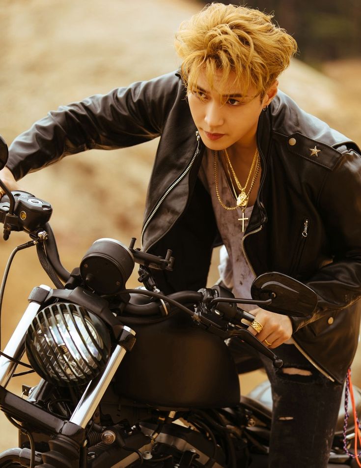 a young man sitting on top of a motorcycle wearing a black jacket and gold necklace