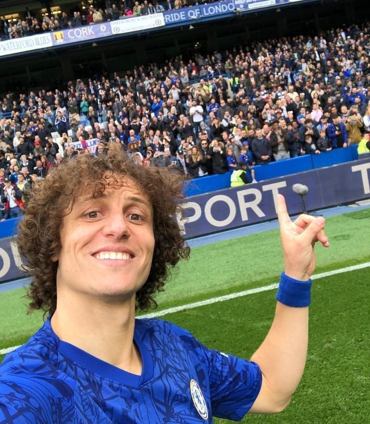 a man with curly hair standing in front of a crowd at a soccer game pointing to the sky