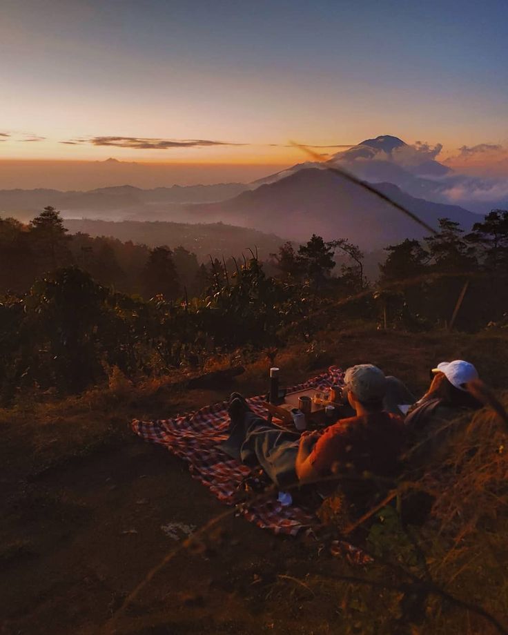 two people are sitting on a blanket in the mountains at sunset, with one person taking a photo