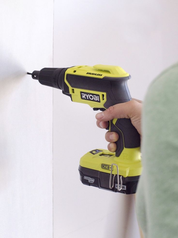 a person holding a cordless drill in their right hand, with a white wall behind them