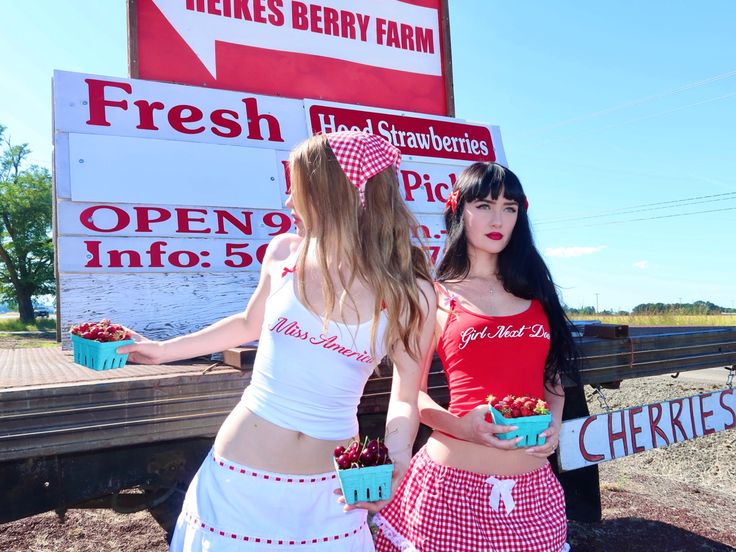 two women dressed in red and white standing next to a sign with cherries on it