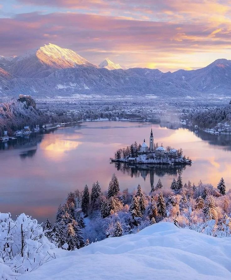 a lake surrounded by snow covered mountains with a church in the middle and trees around it