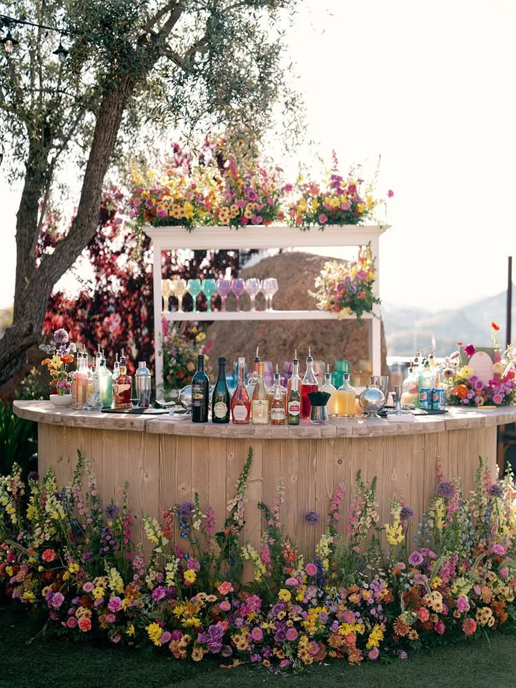 an outdoor bar with lots of bottles and glasses on the top, surrounded by flowers