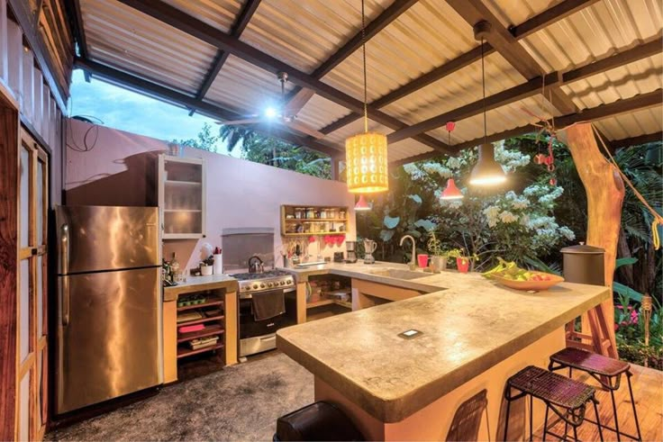 an outdoor kitchen with stainless steel appliances and counter tops is lit by pendant lights hanging from the ceiling