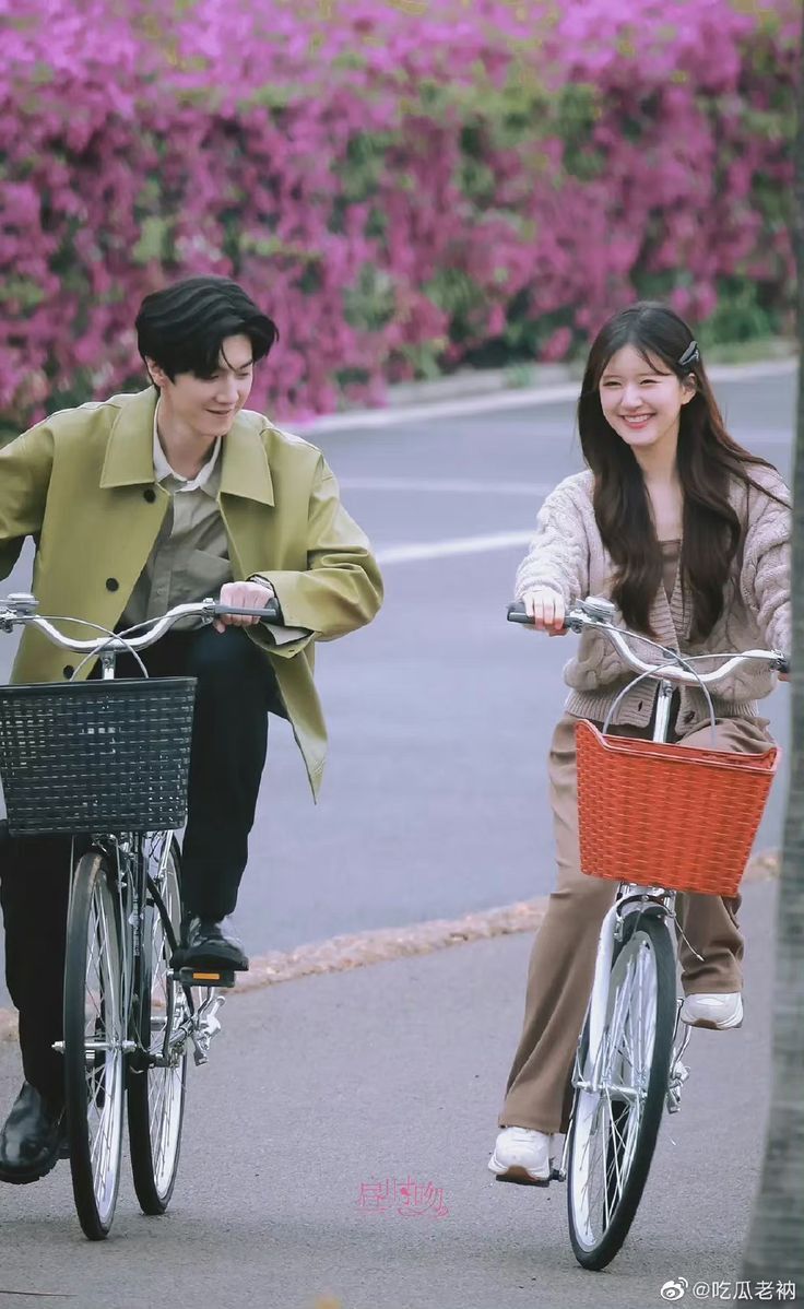 a man and woman riding bikes down the street together with flowers in the back ground