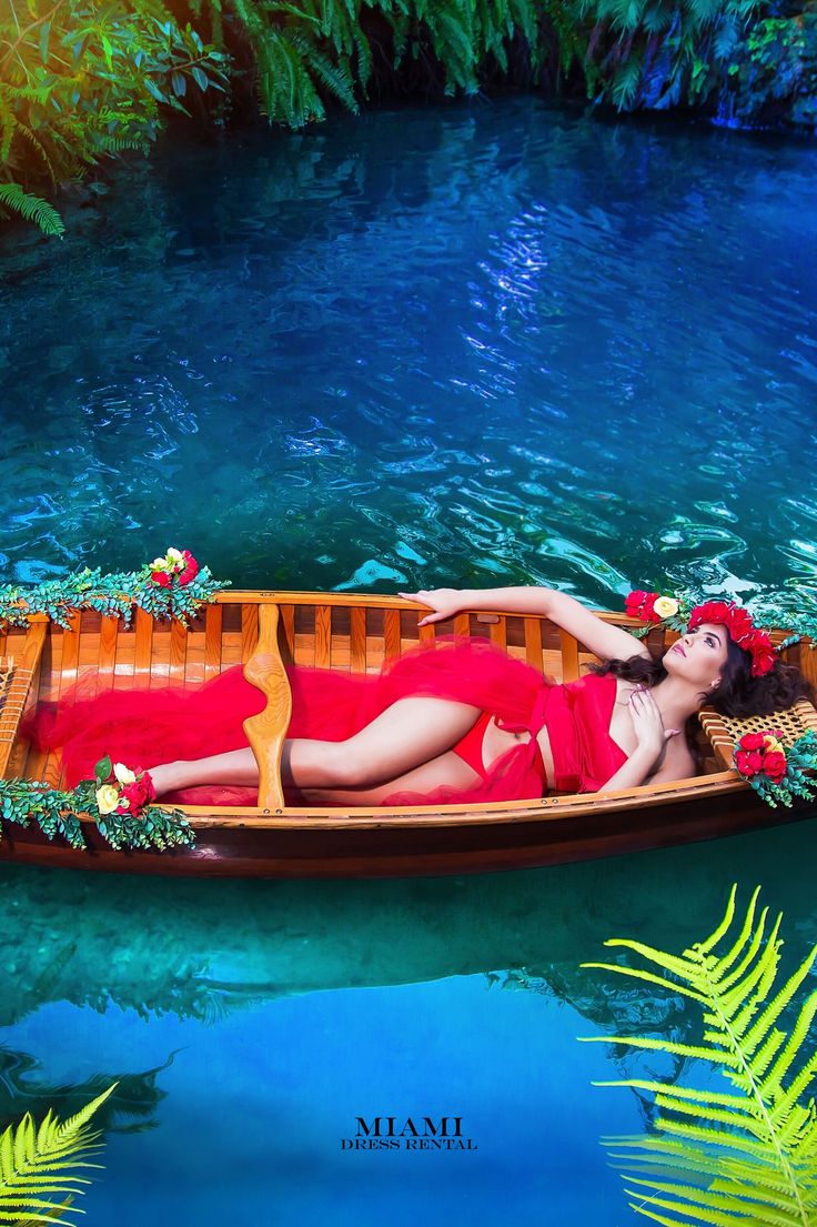 a woman in a red dress laying on a boat floating down a river with palm trees