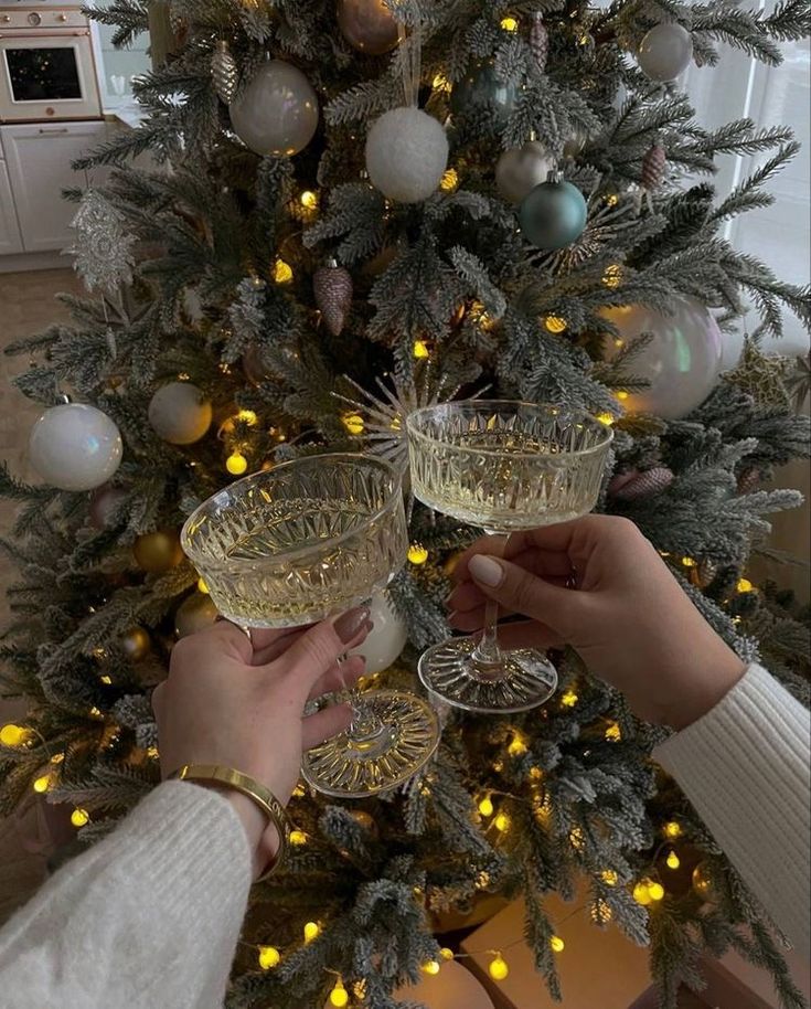 two people toasting champagne glasses in front of a christmas tree with lights on it